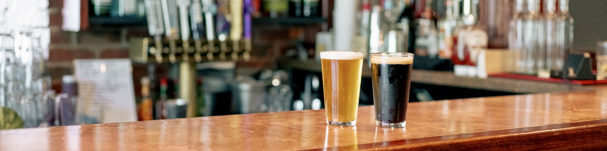 A wooden bar counter with a yellow beer and a dark beer, with a background of various liquor bottles and taps for draft beer.