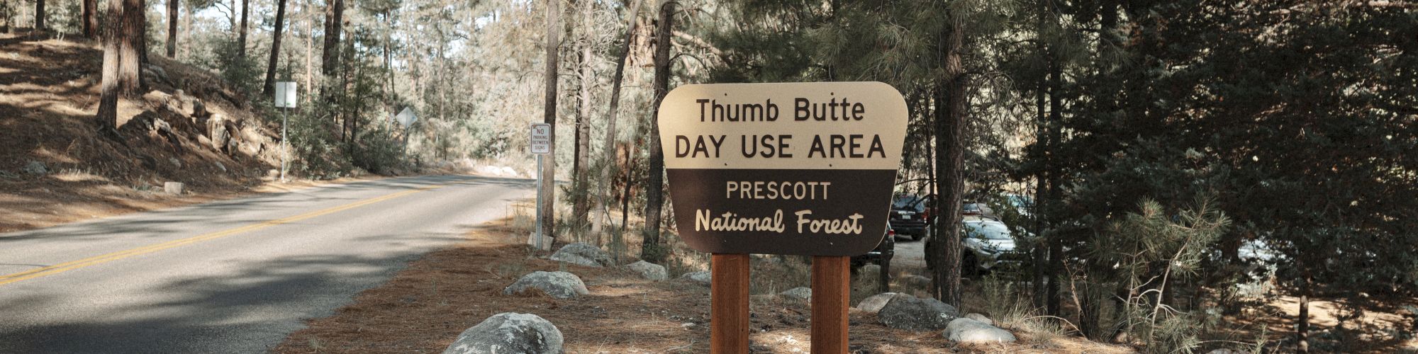 A sign for Thumb Butte Day Use Area in Prescott National Forest, stands by a road surrounded by trees and rocks in a forested area.