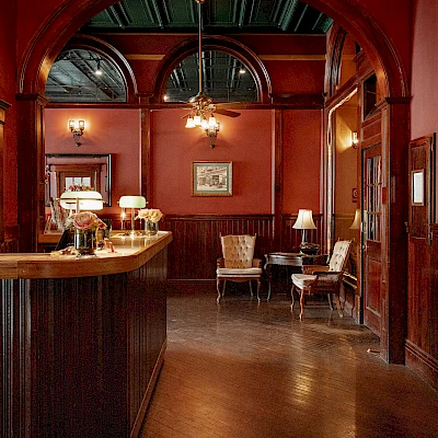 A cozy reception area with a wooden desk, elegant chairs, vintage decor, ambient lighting, and framed pictures on a red wall, creating a warm atmosphere.