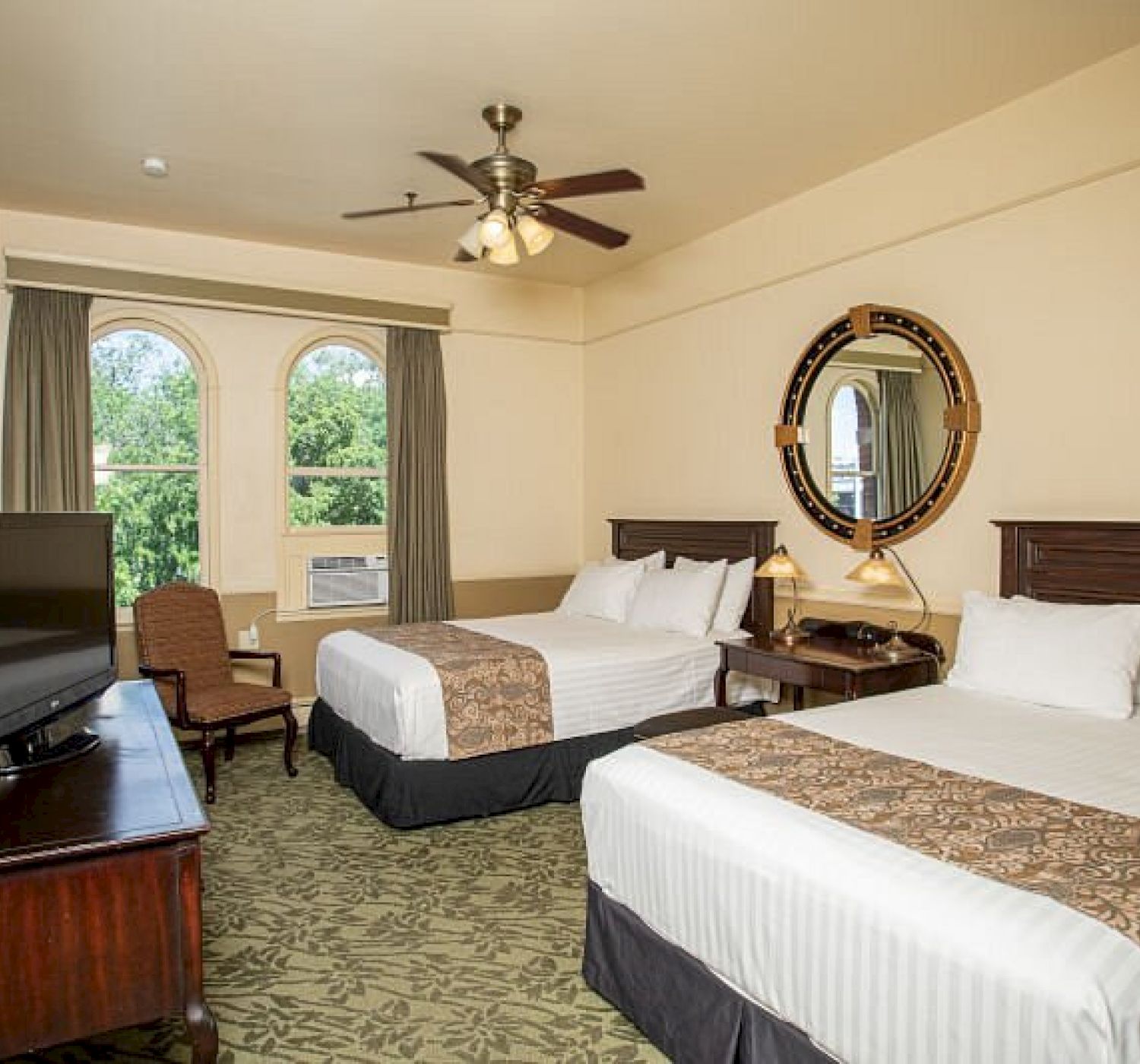 A hotel room with two beds, a TV, a ceiling fan, a chair, a decorative mirror, and large windows with curtains showing trees outside the window.