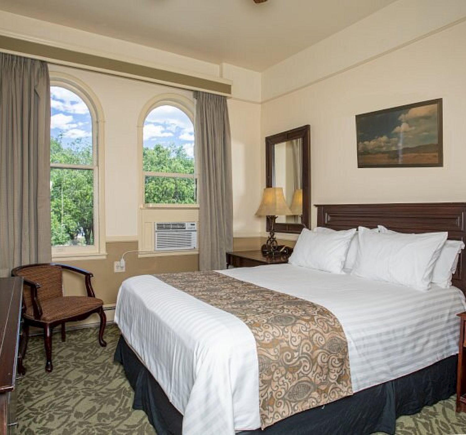 The image shows a cozy hotel room with a double bed, bedside tables, a TV, a chair, and two windows letting in natural light.