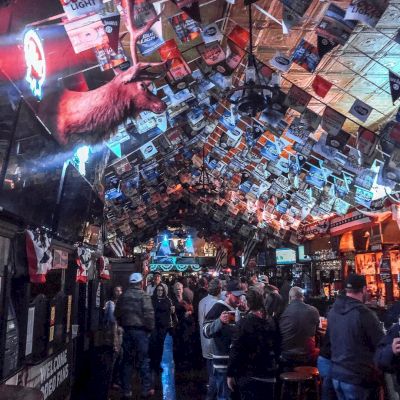 The image shows a crowded bar with a ceiling covered in dollar bills, various neon signs, and a mounted deer head. Patrons are socializing and drinking.