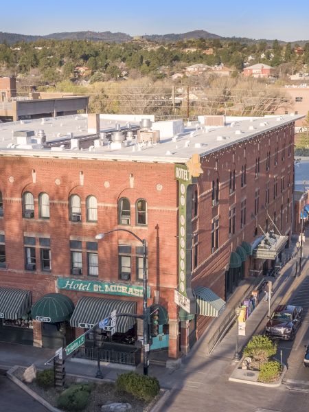 Aerial view of a brick building on a street corner with green awnings, parked cars, and a mountainous background on a sunny day.