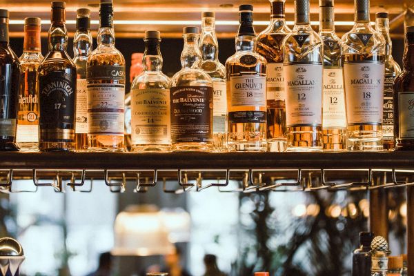 The image shows shelves stocked with various bottles of liquor and spirits in a bar setting, with a warm, glowing ambiance in the background.