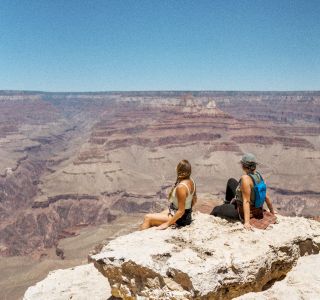 Two people are sitting on a rock ledge overlooking a vast canyon under a clear blue sky. The expansive landscape stretches into the distance.