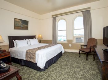 The image shows a hotel room with a double bed, nightstands with lamps, an armchair, a TV on a dresser, and large windows with curtains.