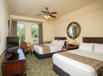 A hotel room with two double beds, a TV, chair, a round mirror, two large windows with green curtains, and a ceiling fan ending the sentence.