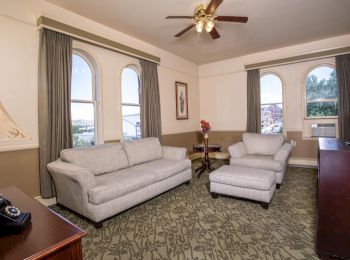 A cozy living room with a sofa, armchair, ottoman, coffee table, lamp, and ceiling fan. Arched windows offer views outside the room.