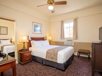 A cozy hotel room with a double bed, bedside tables with lamps, a TV, a ceiling fan, and an adjoining room with another bed.