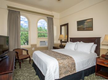 A hotel room with a double bed, a TV on a wooden desk, arched windows with curtains, a chair, lamps, and a wall painting above the bed.