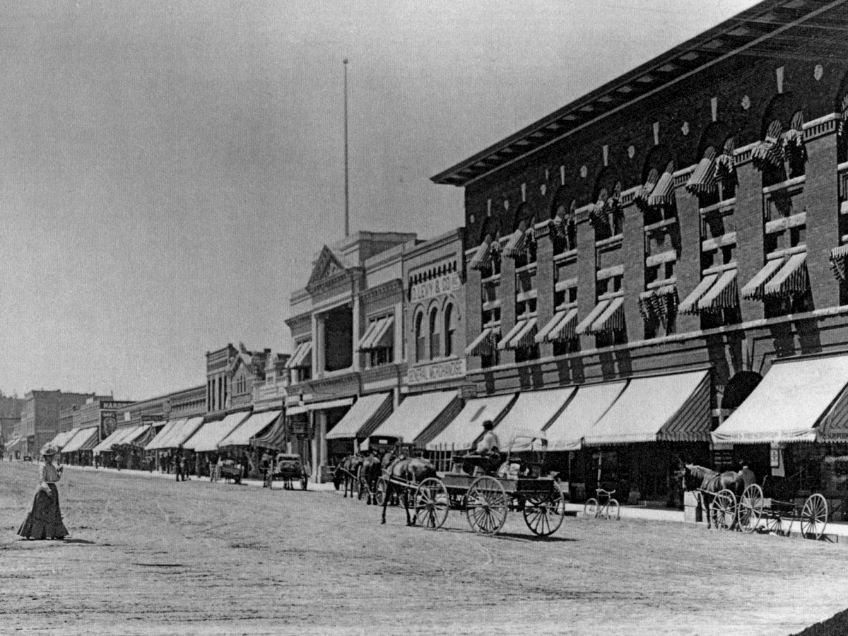 A historic street scene with horse-drawn carriages, buildings with awnings, and a person standing in the foreground.