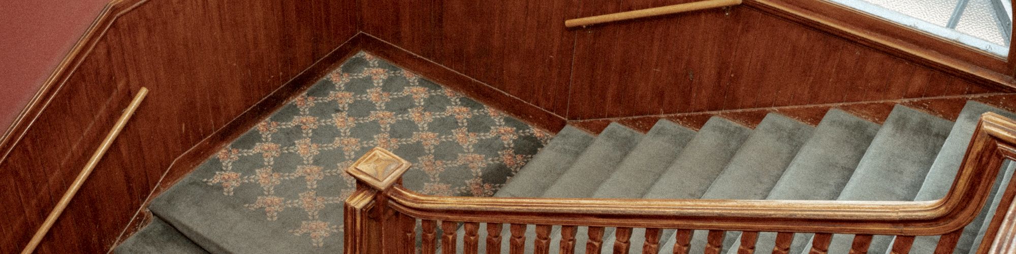 The image shows a wooden staircase with carpeted steps and a partially visible arched window. The wall is paneled and painted in warm tones.