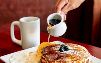 A hand pouring syrup over a stack of pancakes with bacon and a slice of orange on a plate, next to a mug on a wooden table.