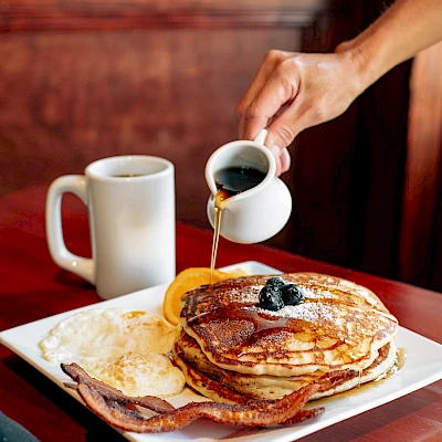 A hand pouring syrup over a stack of pancakes with bacon and a slice of orange on a plate, next to a mug on a wooden table.