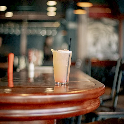 A glass filled with a light-colored beverage sits on a wooden bar counter with stools nearby in a dimly lit setting.