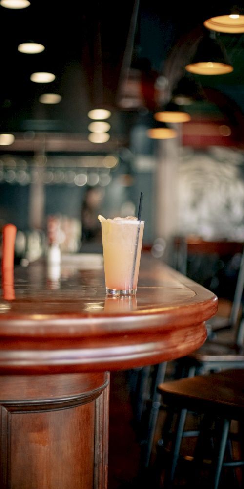 A glass filled with a light-colored beverage sits on a wooden bar counter with stools nearby in a dimly lit setting.