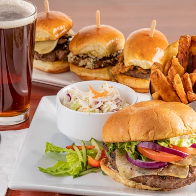 The image shows a meal with a burger, fries, coleslaw, three mini sliders in the background, and a glass of dark beer on a table.