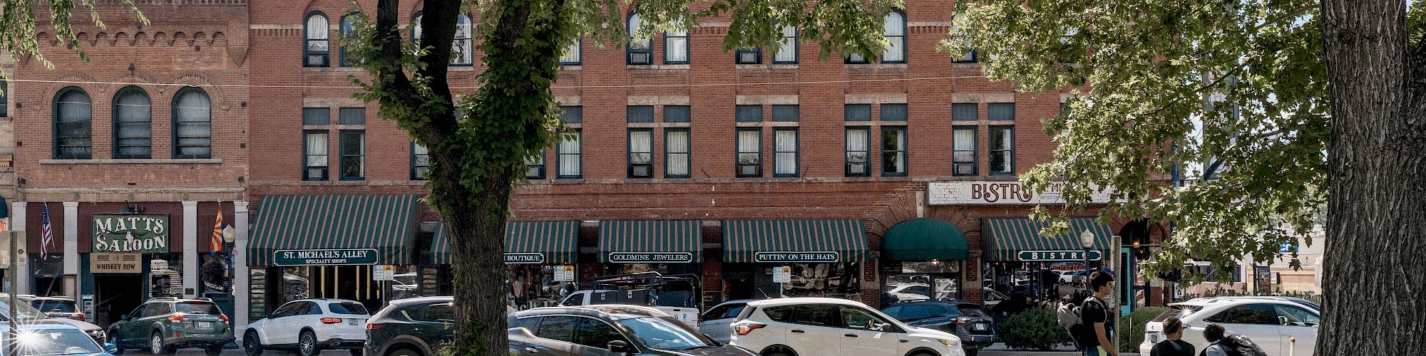 A street view shows parked cars, historical brick buildings, shops with green awnings, pedestrians, and trees in the foreground ending the sentence.