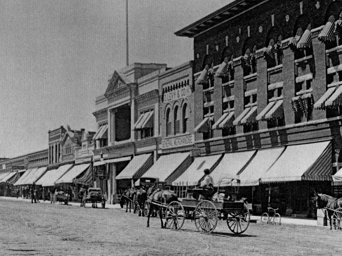 A historic street scene with horse-drawn carriages, storefronts with awnings, and a person standing on the dirt road.