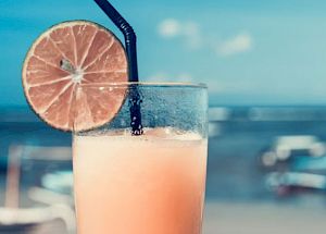 A refreshing beverage in a glass with a black straw and lime slice garnishment is being held in front of a scenic beach backdrop.