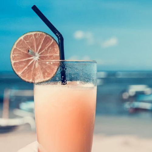 A refreshing beverage in a glass with a black straw and lime slice garnishment is being held in front of a scenic beach backdrop.